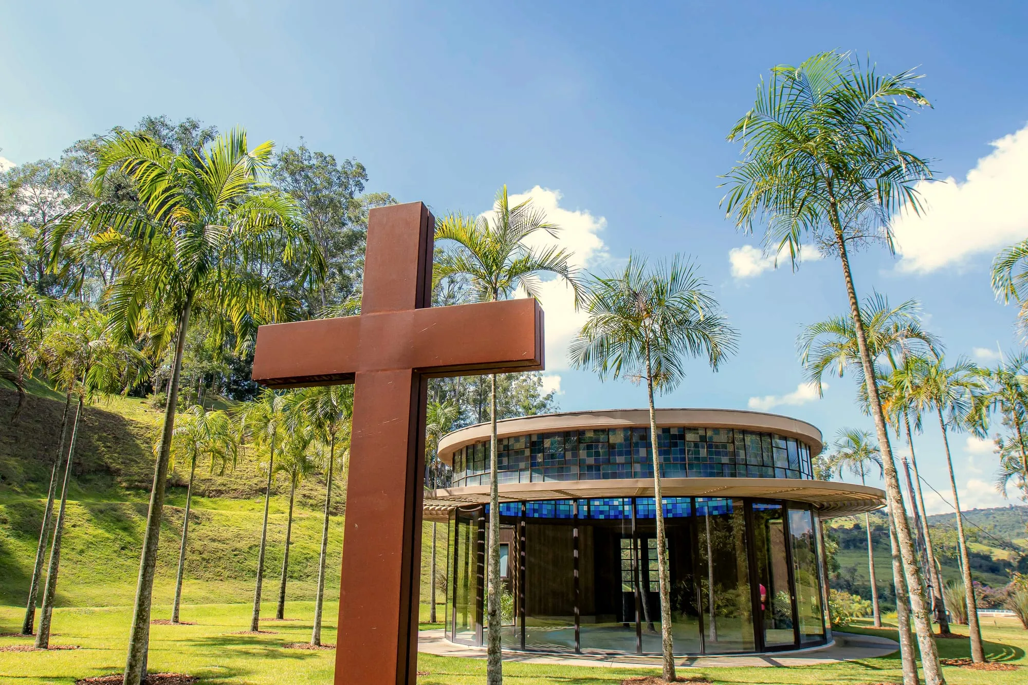 A cruz de aço em primeiro plano e, atrás, a capela redonda de vidro entre palmeiras imperiais
